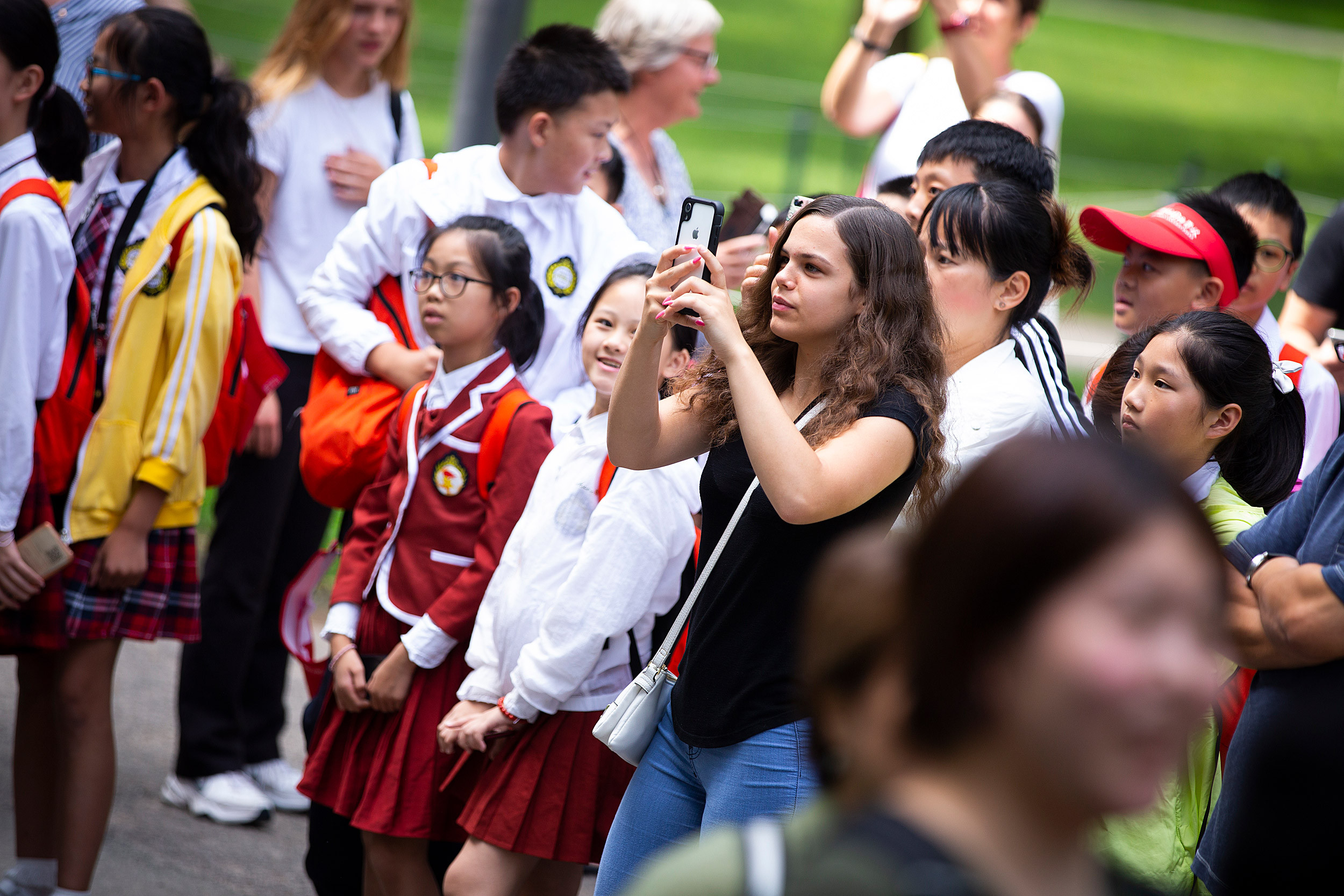 Amidst a crowd of onlookers, a visitor holds up her cellphone to capture a photo of the John Harvard statue, documenting their visit.