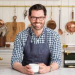John Kanell in his kitchen holding a coffee cup, showcasing his warm and approachable personality.