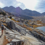 Backpacker hikes toward a rock stair case above an alpine lake on the John Muir Trail on a sunny day