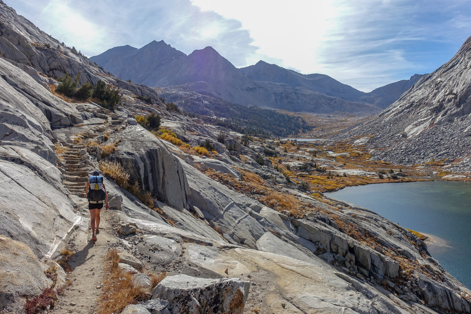 Backpacker hikes toward a rock stair case above an alpine lake on the John Muir Trail on a sunny day